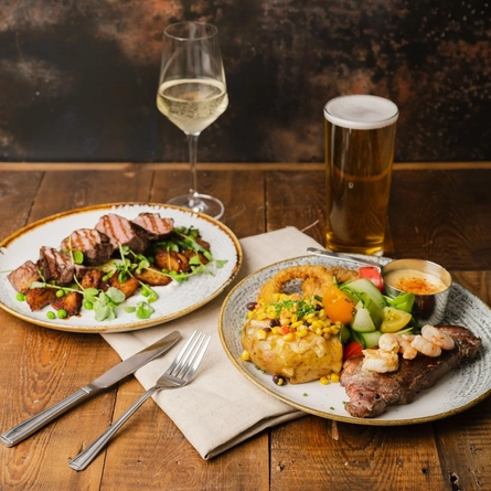 A plate of sliced steak on a bed of roast potatoes; and a plate of steak with prawns, jacket potato, vegetables, onion rings, and a pot of sauce. The plates sit on a cloth napkin on a wooden restaurant table along with cutlery, a glass of white wine, and a glass of beer.