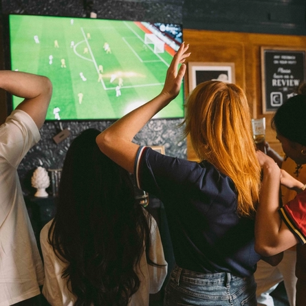 An image showing a group of friends stood watching a football match within the interior seating area at a Greene King venue.