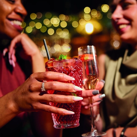 An image of 2 people holding cocktail style drinks celebrating with a festive cheers within the interior of a Greene King Pub venue.