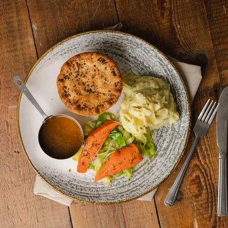 A bird's eye view of a Venison Creamy Green Peppercorn Pie served on a plate with mashed potatoes, vegetables, and a pot of gravy. The plate sits on a cloth napkin on a wooden restaurant table along with a knife and fork.