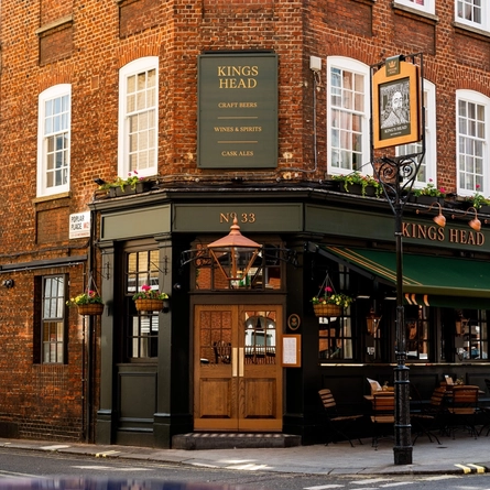 The exterior facade, signage, and seating area at the King's Head in Bayswater, with double wooden doors; a dark green awning hanging over the wooden tables and chairs; flower baskets hanging on the walls; and flower boxes beneath the upper windows.