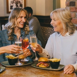 A lifestyle image focusing on 3 friends sat at a table enjoying bottomless burger mains and various drinks, celebrating with a cheers within the interior restaurant seating area at a Greene King venue.