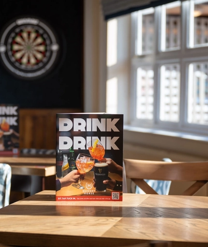 A close up view of wooden tables and chairs in the sunlit interior restaurant seating area of The Weathervane, with a dartboard visible in the background.