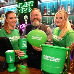 A picture of three Greene King staff members behind a bar with various Macmillan fundraising merchandise.