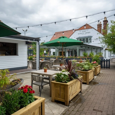 The exterior bar and beer garden seating area at The Kings Arms in Sandford-On-Thames, with wooden planter boxes, shade umbrellas on some of the tables, and string lights overhead.