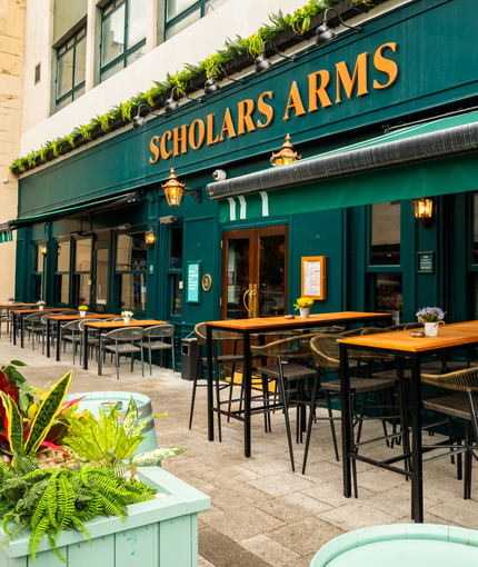 The exterior facade, signage, and seating area at the Scholars Arms in Southampton; with large green awnings hanging above the windows, and wooden planter boxes filled with plants.