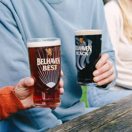 A close up view of two peoples' hands holding glasses of Belhaven Best and Belhaven Black, at a wooden table in a beer garden.