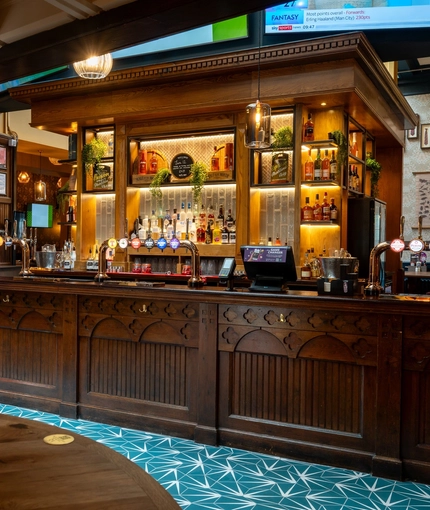 The wood panelled bar inside the Old School House in Aberdeen, with TVs on the wall above it, and bottles of alcoholic spirits on illuminated shelves behind the counter.