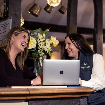Two women sat at a table with a laptop, they are both laughing. 