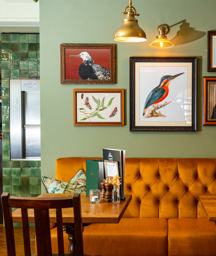 An interior restaurant seating area at The London Apprentice, with a golden yellow velvet booth seat, and framed artwork on the walls above it.