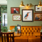 An interior restaurant seating area at The London Apprentice, with a golden yellow velvet booth seat, and framed artwork on the walls above it.