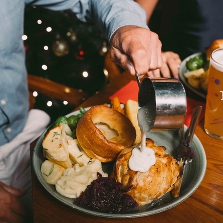 A plate of Christmas dinner including Yorkshire pudding, roast potatoes, mashed potatoes, vegetables, and Brussels sprouts; sitting on a wooden restaurant table. Serviettes and another plate of food also sit on the table, and a person sitting at the table is pouring sauce from a small pot over the plate.