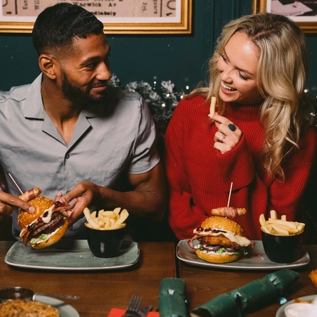 Two people sitting at a restaurant table with plates of burgers and chips in front of them. One person is picking up their burger, the other person is holding a chip, and a Christmas garland lit with fairy lights is visible in the background.