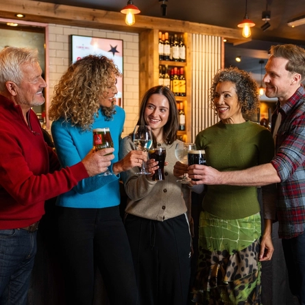 An image of a group of people enjoying drinks and celebrating with a cheers, stood at the bar within a Flaming Grill venue.