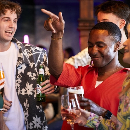 A group of people talking and dancing inside a pub bar area, holding drinks. Some hold glasses of wine, cocktail or beer, one holding a bottle of zero alcohol beer. 