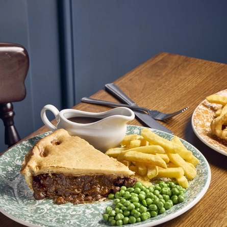 A plate of pie, chips, peas, and a jug of gravy, and a plate of fish, chips, and peas, sitting on a wooden table with a knife and fork.