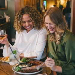 Two women seated at a wooden table, enjoying a meal and drinks.