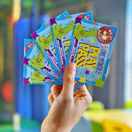 A close up view of a hand holding five Wacky Warehouse One Month Play Pass cards, with a children's indoor play area visible in the background.