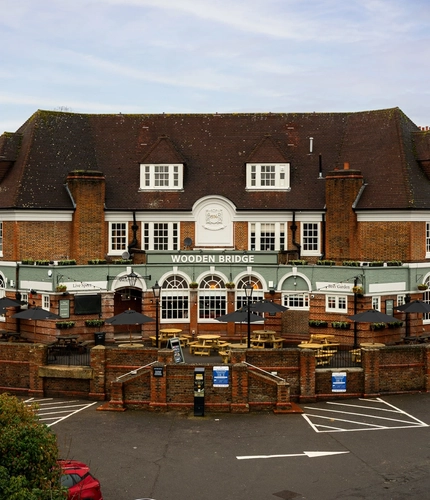 The exterior facade, signage, and beer garden seating area of the Wooden Bridge in Guildford, with wooden picnic tables and shade umbrellas.