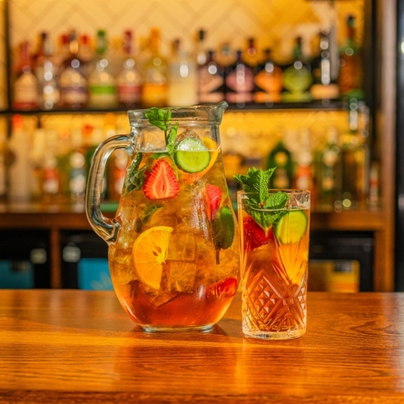 A close up view of a large glass jug filled with Pimm's cocktail, ice, mint, and slices of orange, strawberry, and cucumber. The jug stands on the bar inside a pub, along with a Pimm's cocktail served in a highball glass and garnished in the same way.