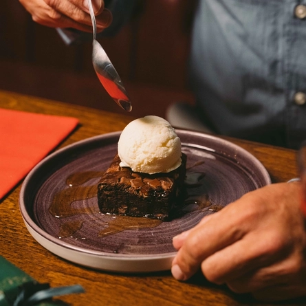 A close up view of a slice of chocolate brownie drizzled with sauce, topped with a scoop of ice cream, and served on a plate. The plate sits on a wooden restaurant table along with a glass of mulled wine, a red serviette, and a green Christmas cracker; and a person at the table is holding a spoon above the plate.