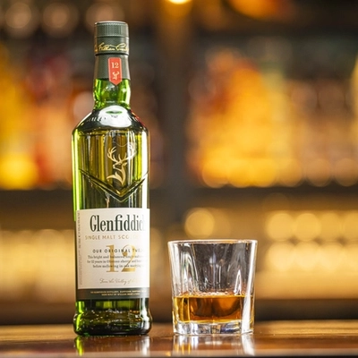 A close up view of a bottle of Glenfiddich standing on table, along with a small amount of the whisky served in a whiskey glass.