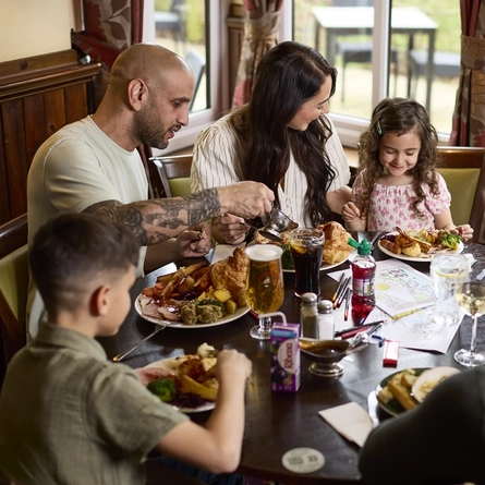 Three adults and three children sitting at a restaurant table in a Farmhouse Inns venue, with drinks, plates of roast dinner, colouring pages and pencils in front of them.