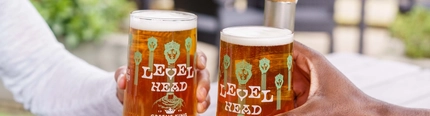 A close up image focusing on 2 pints of Level Head being held within an exterior beer garden seating area.