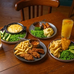 A wooden restaurant table laden with plates of children's meals including Chicken Tenders with fries and baked beans, Fish & Chips with peas and vegetable sticks, Vegan Sausages with peas, mashed potatoes and gravy, and Mac & Cheese with peas and vegetable sticks. Two glasses of orange juice also sit on the table.