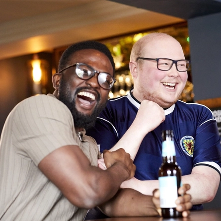 Inside a Greene King pub during a Six Nations match day, two people sit closely together celebrating at a wooden table. One person is wearing a dark rugby shirt with a national team crest, holding a beer bottle on the table. The other person sits beside them, resting an arm on the table in a friendly, supportive posture. In front of them is a tall glass of pale lager, and the background shows softly lit pub décor, including shelves of bottles and warm lighting. The scene captures the sociable, match‑day atmosphere of fans gathering to watch the Six Nations tournament.