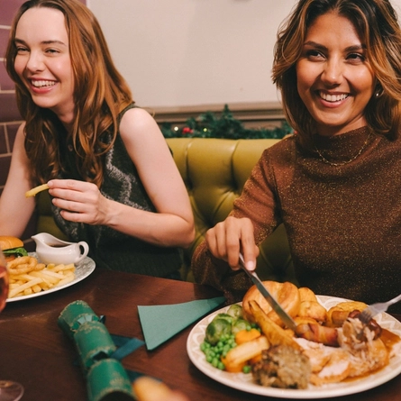 An image of a group of friends sat at a table enjoying festive main dishes and various drinks within the interior restaurant seating area at a Proper Local venue.