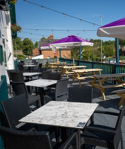 The exterior seating area on the deck of the Rudds Arms; with wooden picnic tables, shade umbrellas, and string lights overhead.
