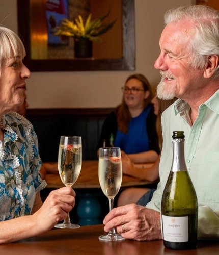 A couple seated at a restaurant table with champagne glasses and a bottle of Prosecco between them.