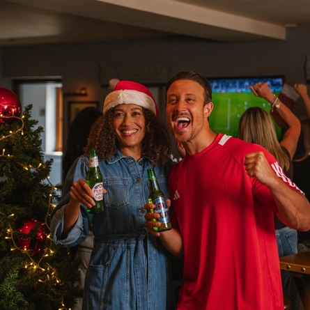 A Christmas-decorated pub with a large tree adorned with red baubles, ribbon and warm fairy lights. Two people stand beside the tree holding bottled beers, one wearing a red-and-white Santa hat and the other a red sports shirt. A television in the background displays a football match while several other patrons watch with raised arms. Wooden tables and chairs are visible throughout the venue, with a plate of onion rings on a nearby table.