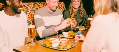 Three people sit in a festive pub booth decorated with Christmas garlands and red baubles. A wooden table is spread with sharing dishes including pigs in blankets, roast potatoes, dipping sauces and burgers, alongside glasses of lager, stout and red wine. One person takes a large bite from a stacked burger while the others enjoy food and drinks in a cosy Christmas pub setting.