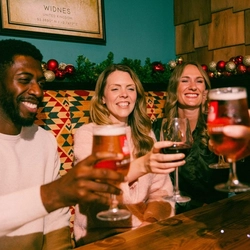 Four people sit together in a cosy pub booth decorated with Christmas garlands and red and gold baubles. The group raises glasses of lager and red wine in a toast over a wooden table, while festive drinks and seasonal décor create a warm and sociable Christmas pub atmosphere.