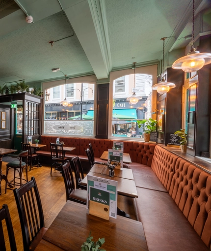 An interior restaurant seating area at The Plough in Bloomsbury, with a long, upholstered seat running along two sides of the room, and a TV on the wall.