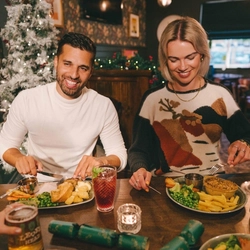Four people sitting at a restaurant table with plates of food in front of them. Glasses of Level Head IPA, Guinness and a cocktail also sit on the table and one person is raising a glass of champagne.