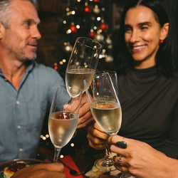 A close up view of four glasses of sparkling wine being clinked together by a group of people sitting around a restaurant table laden with plates of food. A Christmas tree decorated in red and gold and lit with fairy lights is visible in the background.