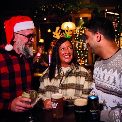 Three people are standing at a tall table with drinks,  in a festive, warmly lit bar. Two of them are wearing Christmas themed headwear.  The background features Christmas decorations, including garlands and lights, as well as hanging glasses above the bar area.