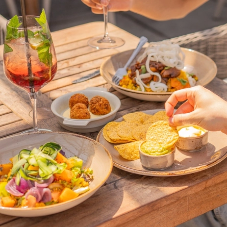 A sharer plate of Nachos served with two pots of dipping sauce sitting on a sunlit wooden table in a beer garden, along with cutlery, three other dishes of food, and a cocktail served in a wine glass. Two people are sitting at the table, one of them holding another cocktail, and the other dipping one of the nachos into a pot of sauce.