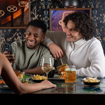 Three people sit around a restaurant table. One person leans toward another while showing something on a phone. The person beside them has an arm around their shoulders. Across the table, another person sits facing them. The table is filled with dishes including fries, salad, and a main dish, along with drinks such as a glass of white wine and a pint of beer.