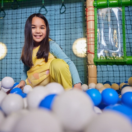 A child smiling at the camera in a ball pit in an indoor play area.