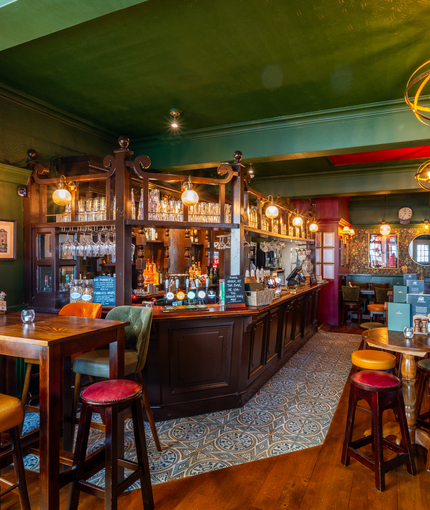 The interior restaurant seating area and wood panelled bar at the Two Chairmen, with upholstered chairs and bar stool seating, wooden flooring, and framed artwork on the floral wallpapered walls.