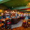 The interior restaurant seating area and wood panelled bar at the Two Chairmen, with upholstered chairs and bar stool seating, wooden flooring, and framed artwork on the floral wallpapered walls.