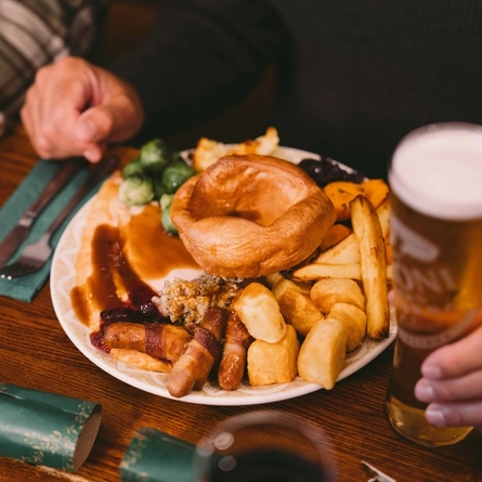 Festive roast dinner with Yorkshire pudding, pigs in blankets, roast potatoes, Brussels sprouts and gravy, served on a wooden pub table beside a pint of beer and green Christmas crackers.
