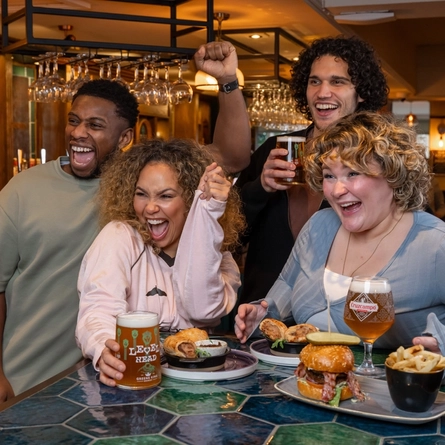 Four people stand and sit around a bar-height table in a busy pub. The table is covered with food, including sliders, fries, dips, and small plates. Several drinks are visible, such as a pint of beer, a canned drink, and another beer in a stemmed glass. One person standing behind the table holds a pint.