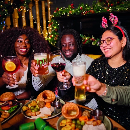 A group of people celebrating a festive Cheers while enjoying various main meals and drinks within the interior restaurant and seating area of Greene King Pub venue.