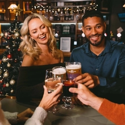 Four people at a table in a bar, clinking together glasses of beer, white wine, and mulled wine. A Christmas tree decorated in red and gold, and lit with fairy lights stands behind the table.