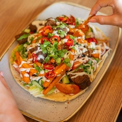 A plated Lamb Kofta on a board on a table, a hand is taking a french fry.